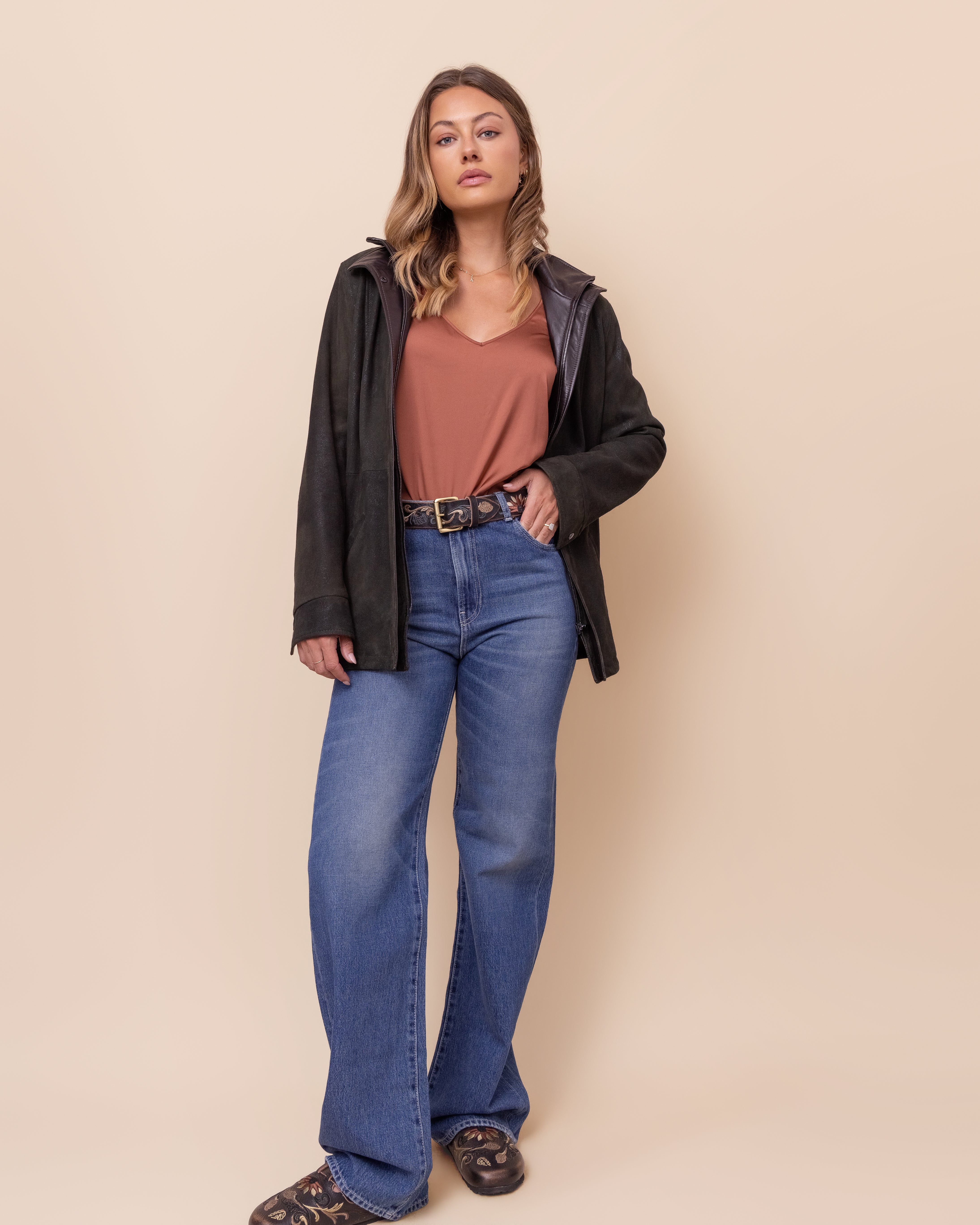 Woman wearing a green leather jacket, brown top, and blue jeans against a beige background