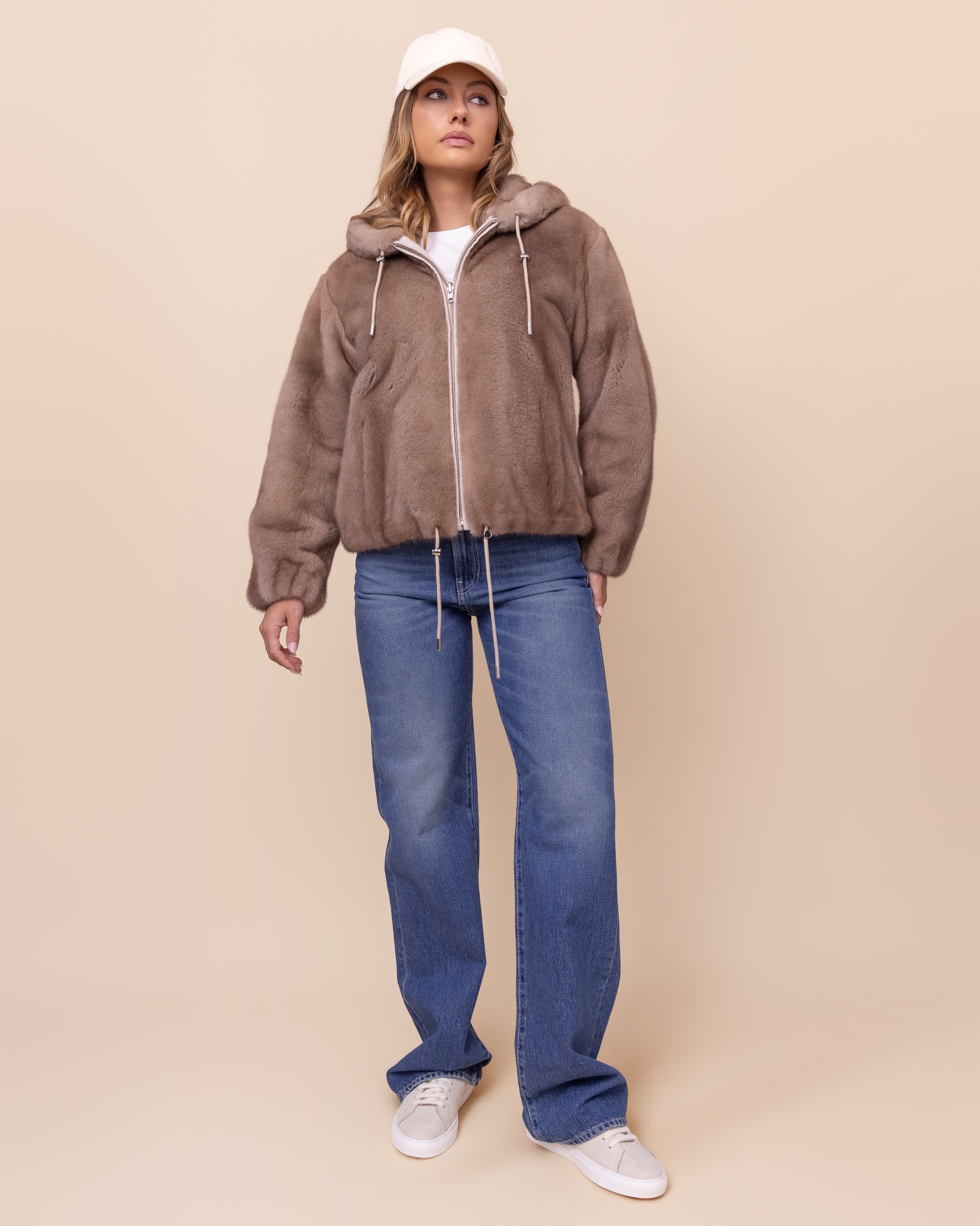 Person wearing a mink jacket, blue jeans, and white sneakers on a beige background