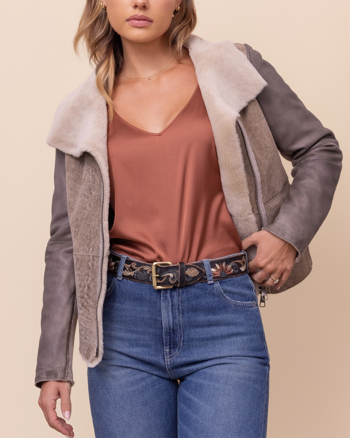 Woman wearing a gray shearling jacket with a cream collar, brown top, and blue jeans on a beige background.