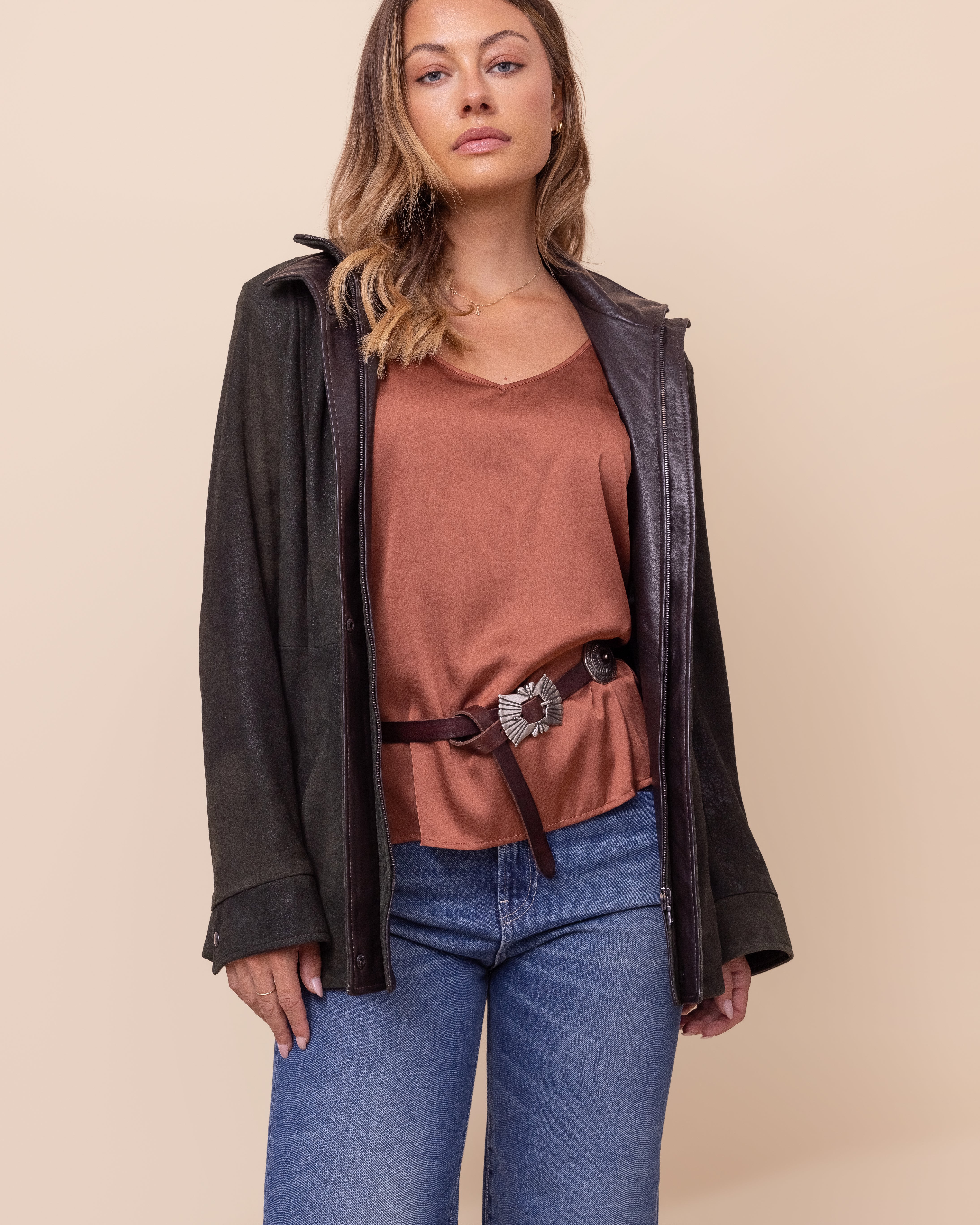 Woman wearing a green and brown leather jacket, rust-colored top, and blue jeans on a beige background