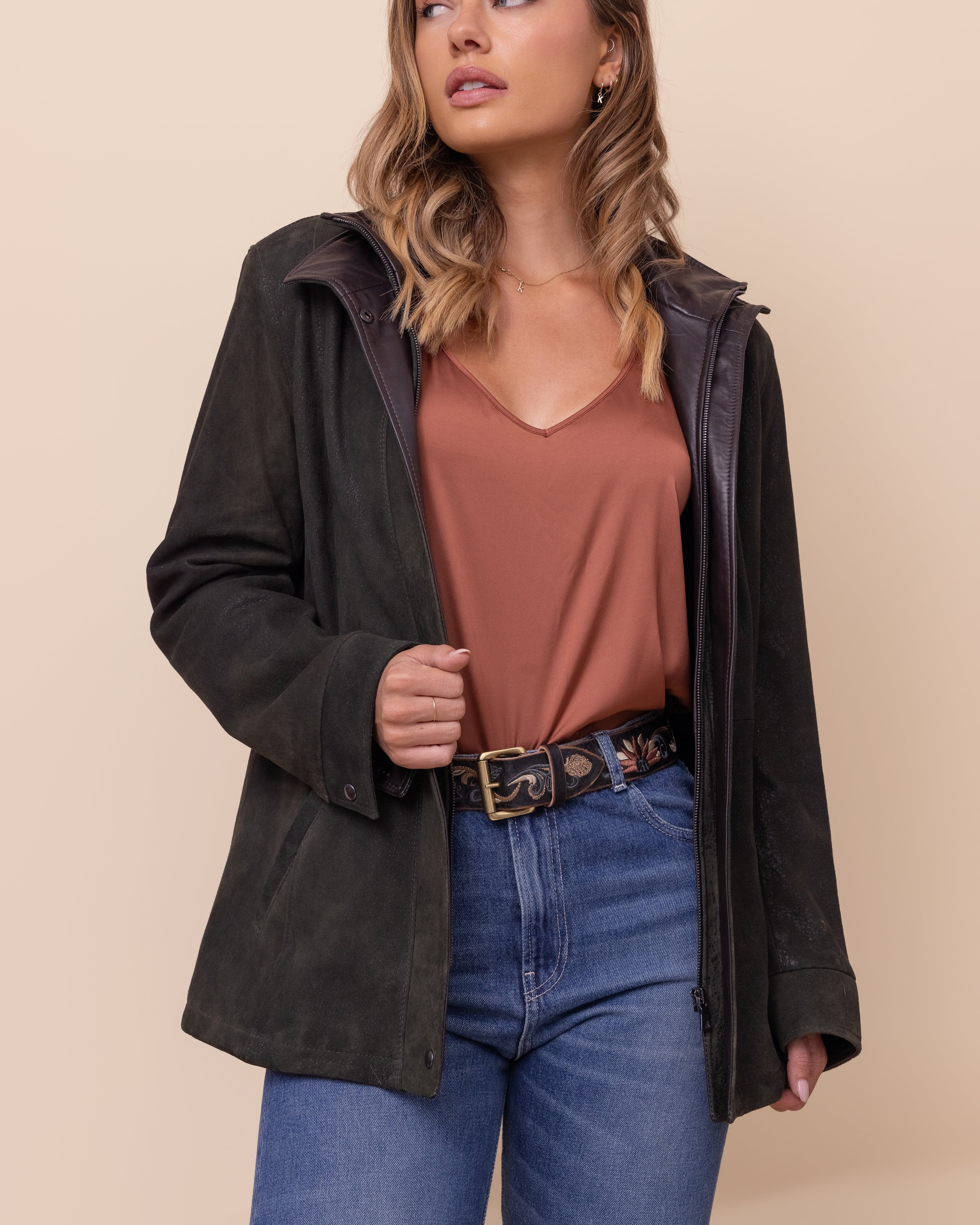 Woman wearing a green jacket, brown top, and blue jeans against a beige background