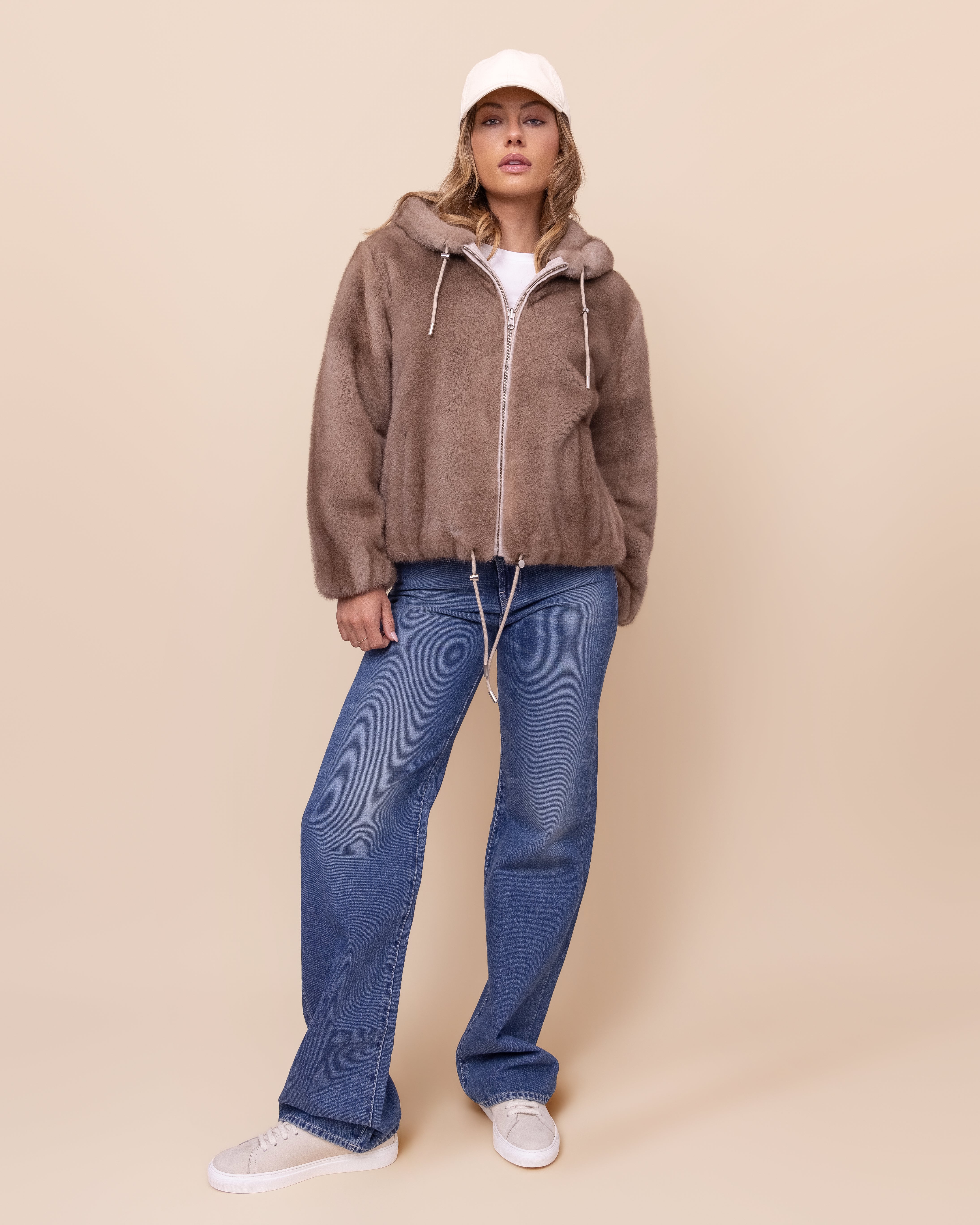 Person wearing a brown mink jacket, blue jeans, and a white cap on a beige background