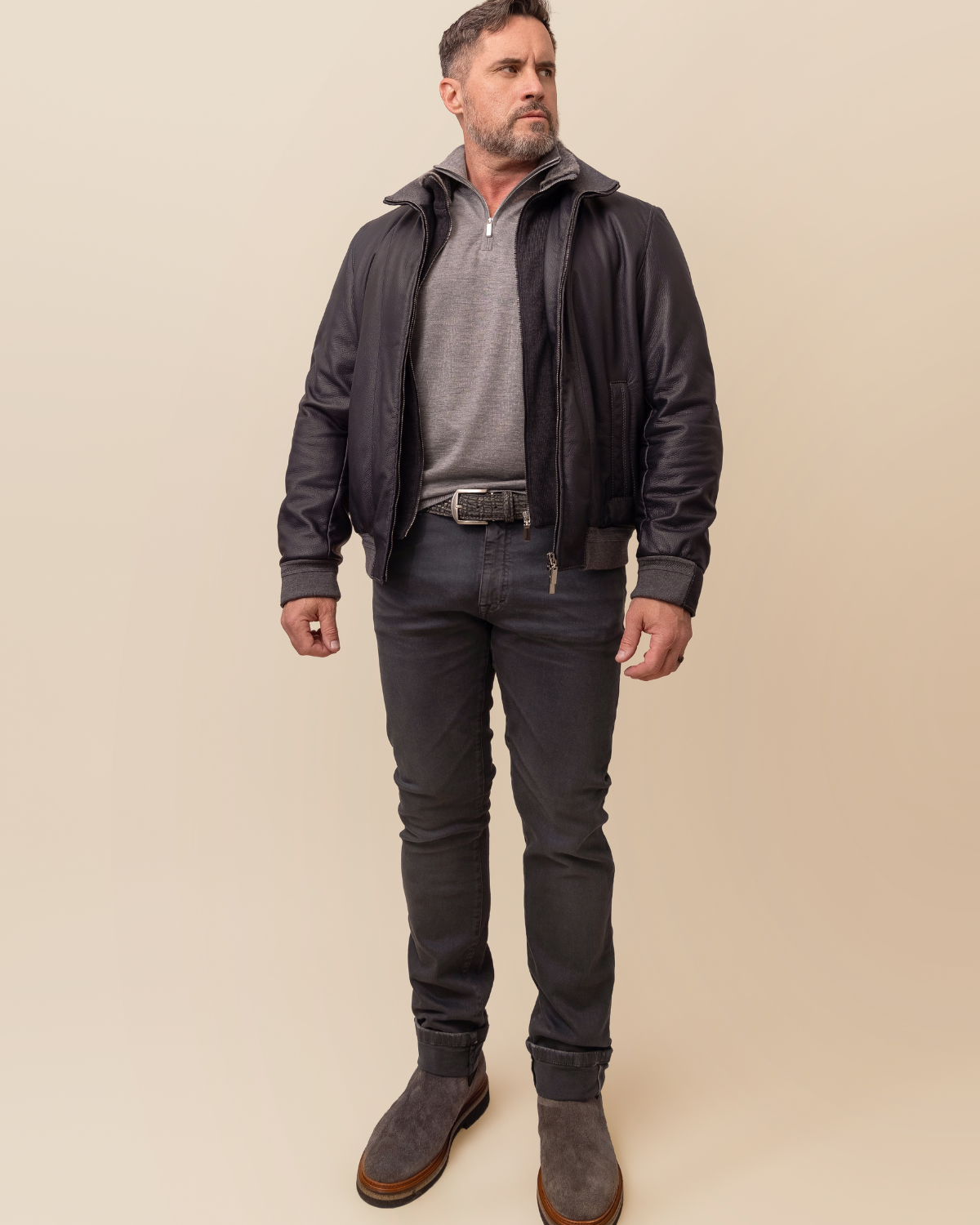 Man wearing a dark jacket, gray shirt, and jeans on a beige background