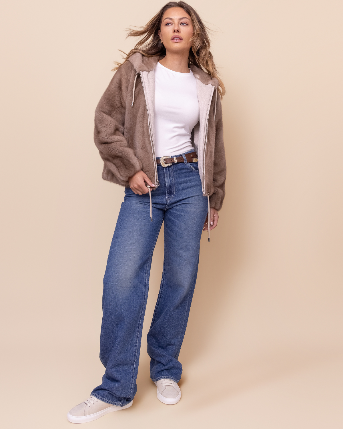 Woman wearing a brown mink jacket, white shirt, blue jeans, and white sneakers on a beige background