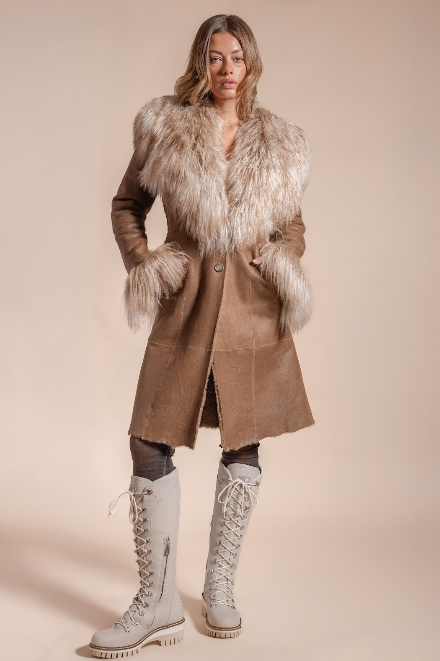 Woman wearing a long brown coat with fur trim and white boots on a beige background