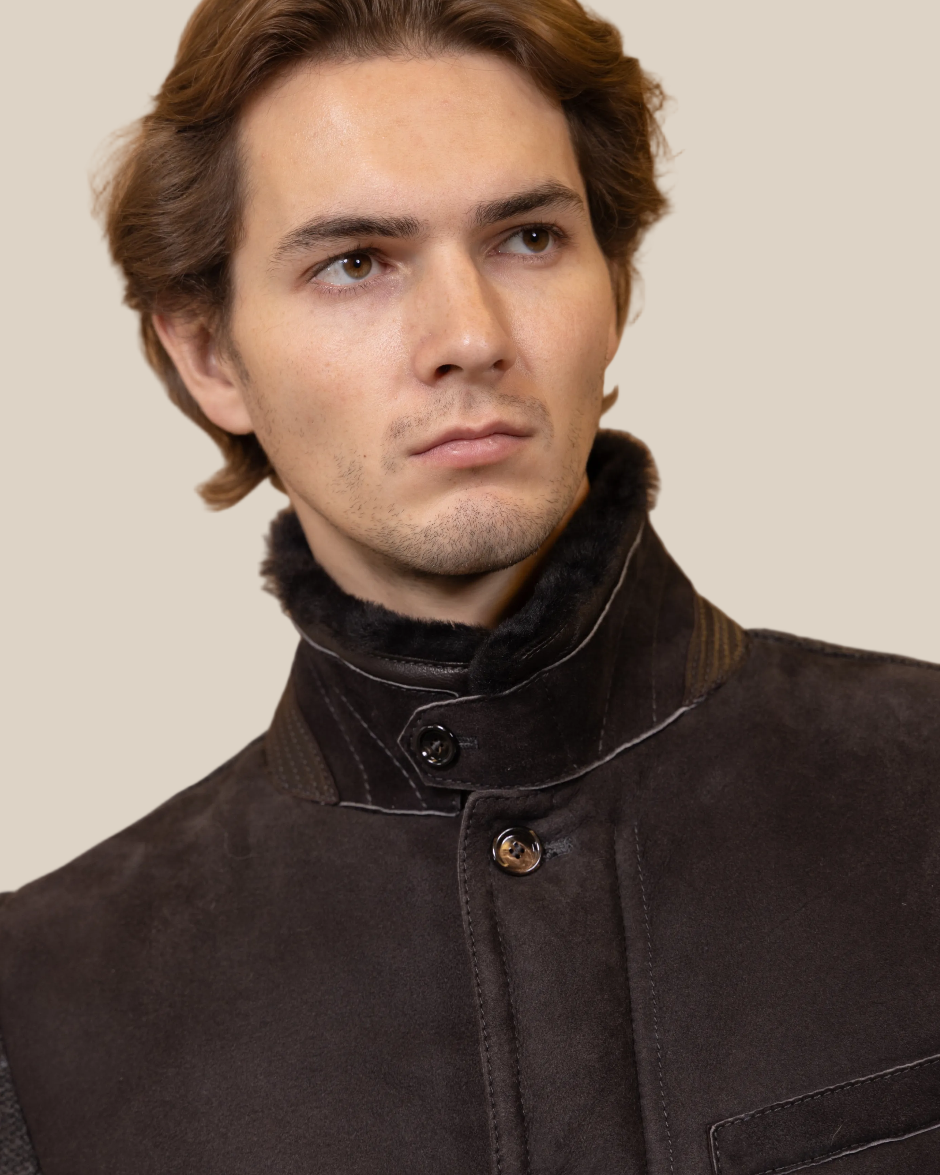 Man wearing a dark brown coat with a high collar against a beige background